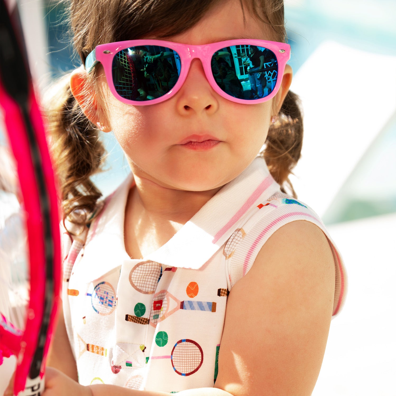 Child wearing pink sunglasses and A cream sleeveless polo dress with a grey and white striped collar, decorated with a whimsical pattern of multi-colored tennis and badminton equipment, by Magnetic Me.