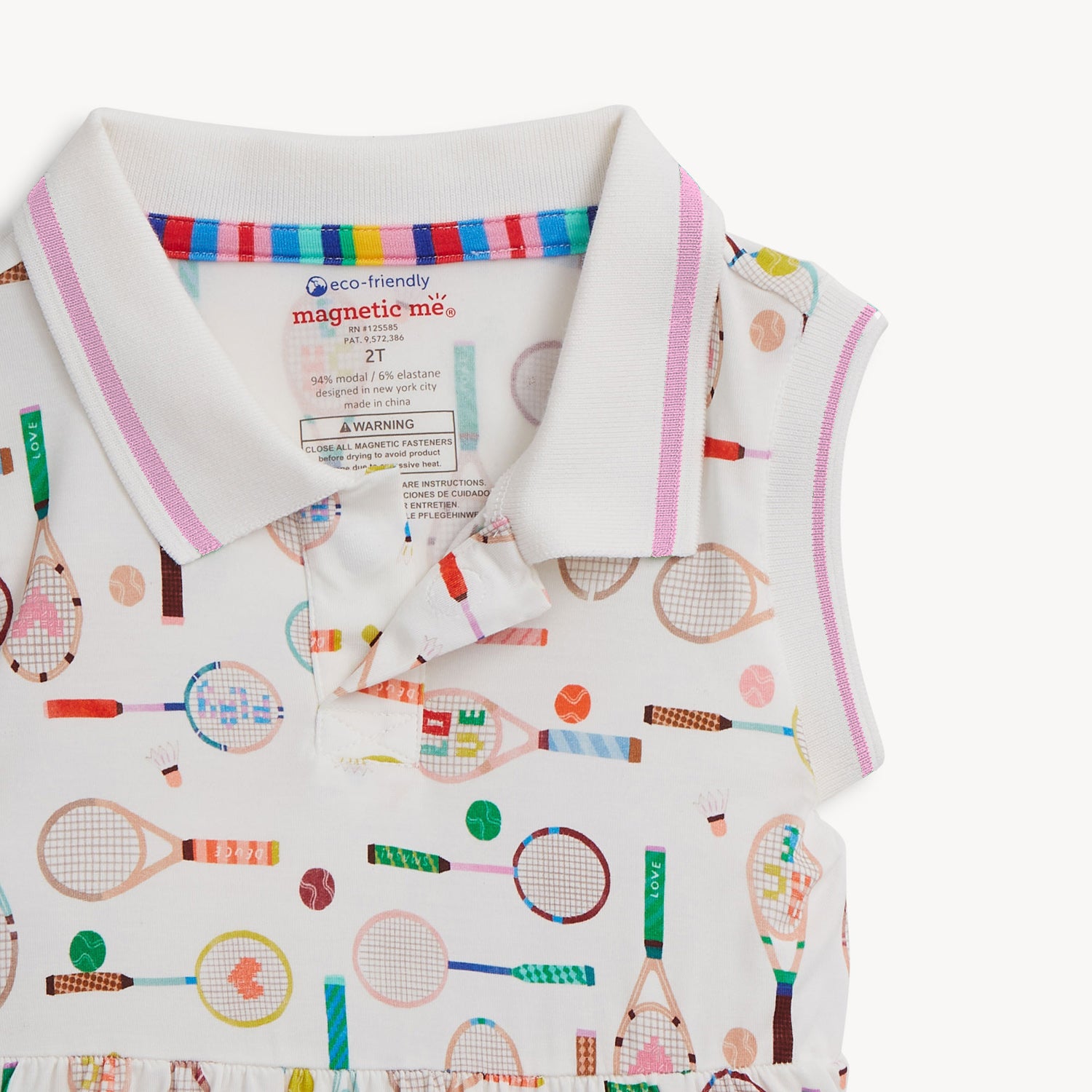Open magnet on the front bottom of the magnet of A cream sleeveless polo dress with a pink and white striped collar, decorated with a whimsical pattern of multi-colored tennis and badminton equipment, by Magnetic Me.