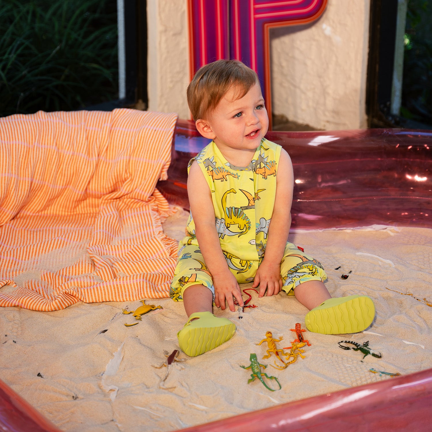 Child playing with toys on a sandy surface wearing A sleeveless one-piece romper in lime green with a vibrant all-over print of prehistoric dinosaurs and flying pterodactyls, by Magnetic Me.
