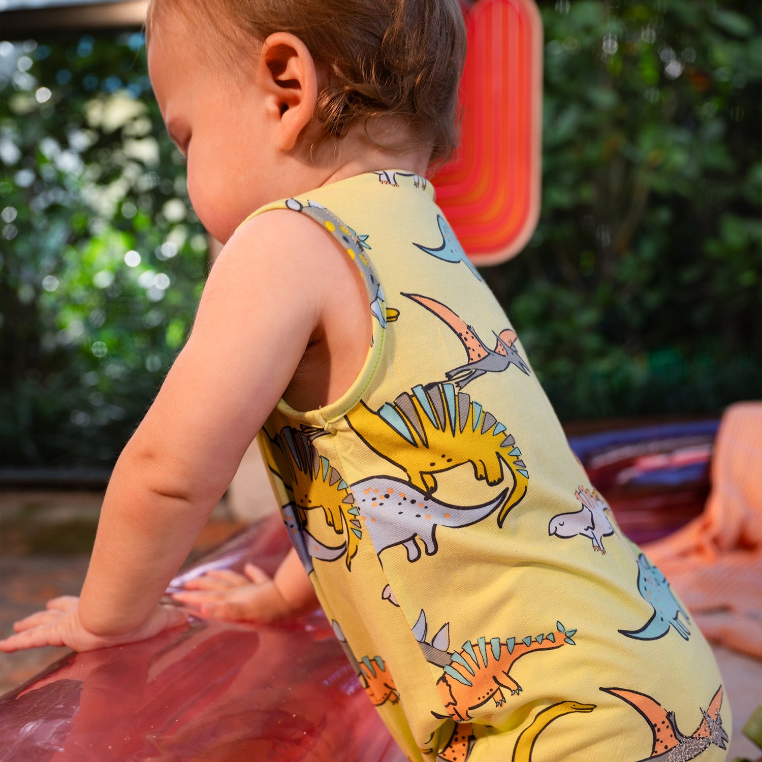 Child leaning on the inflatable pink pool while wearing A sleeveless one-piece romper in lime green with a vibrant all-over print of prehistoric dinosaurs and flying pterodactyls, by Magnetic Me.
