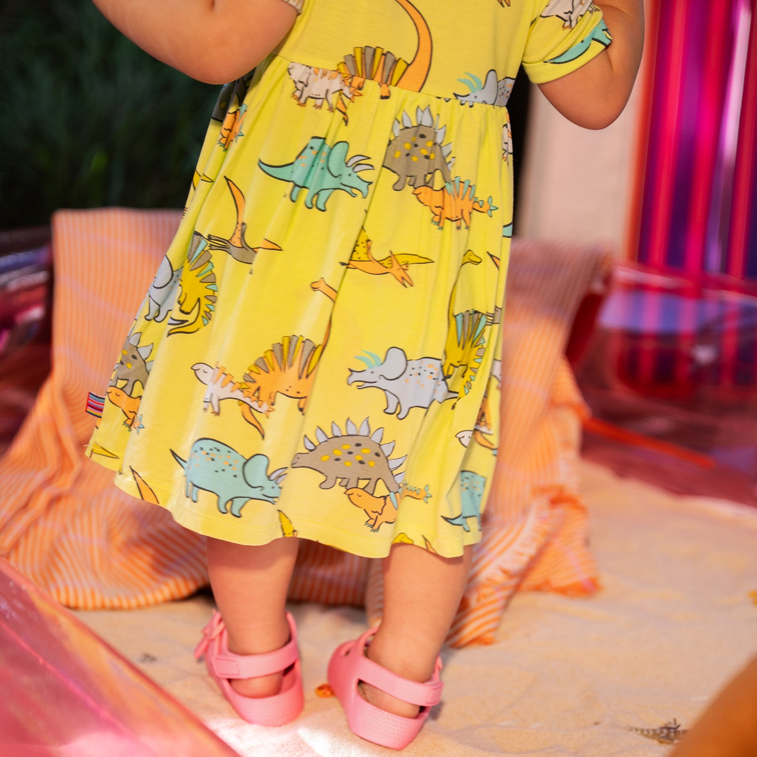 Baby showing the bottom area of A lime green short-sleeve infant dress with a gathered waist, small chest pocket, and an integrated bodysuit featuring a colorful dinosaur pattern, by Magnetic Me.