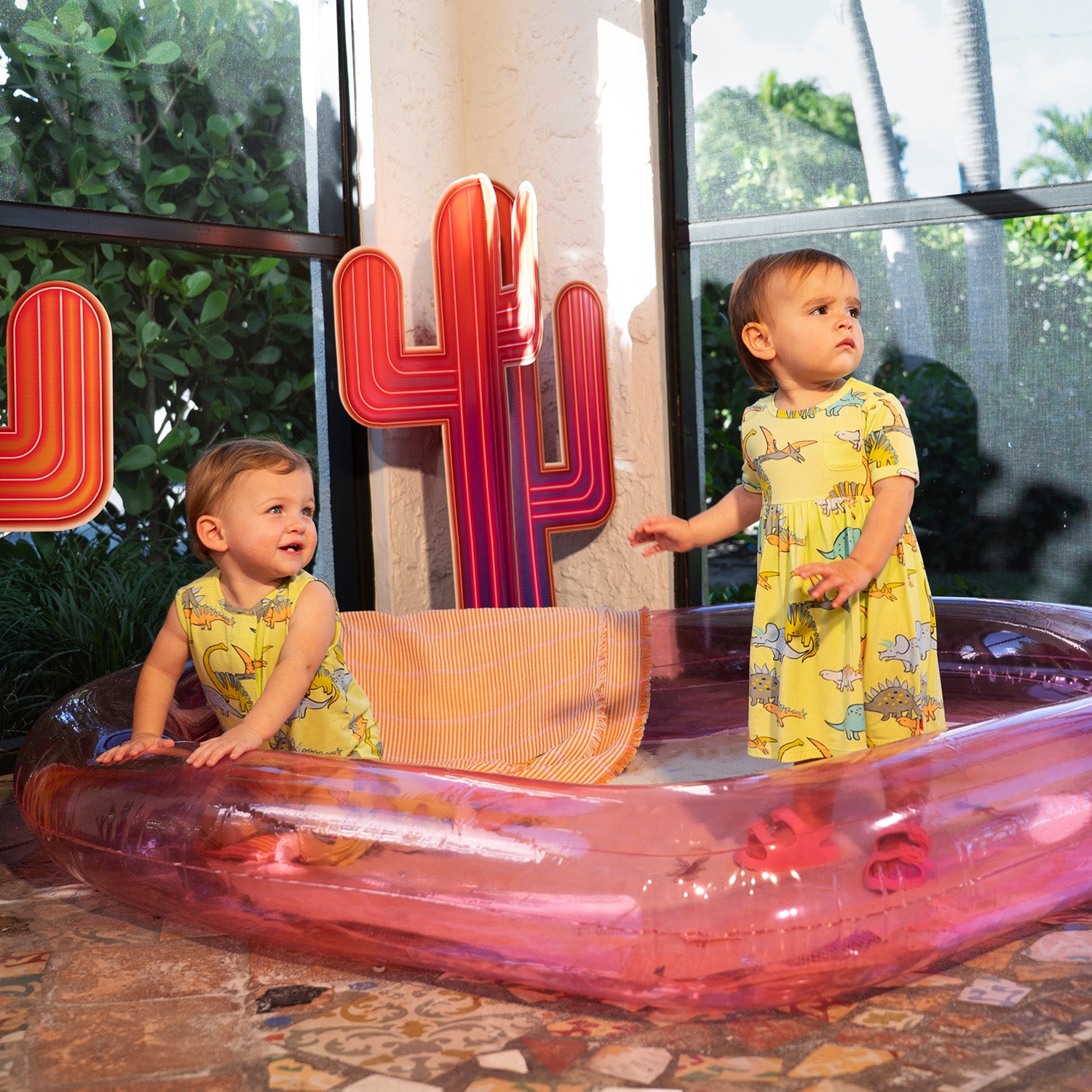 Two children standing on a transparent pink inflatable pool with decorative letters in the background.