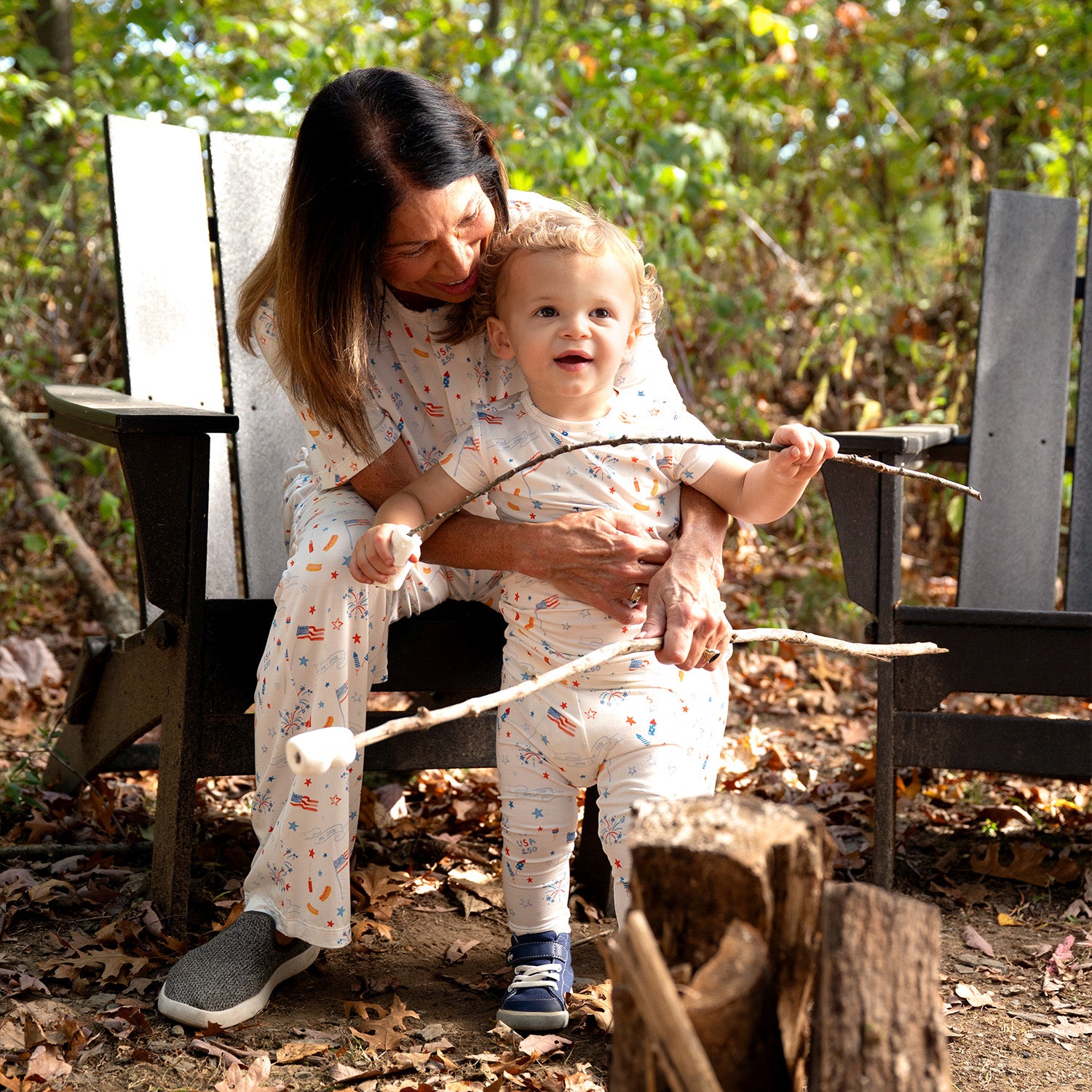 Woman and child sitting on a wooden bench outdoors with sticks wearing A two-piece white pajama sets with a short-sleeve top and shorts featuring a colorful celebratory print of rockets and "1776-2026" banners, by Magnetic Me.
