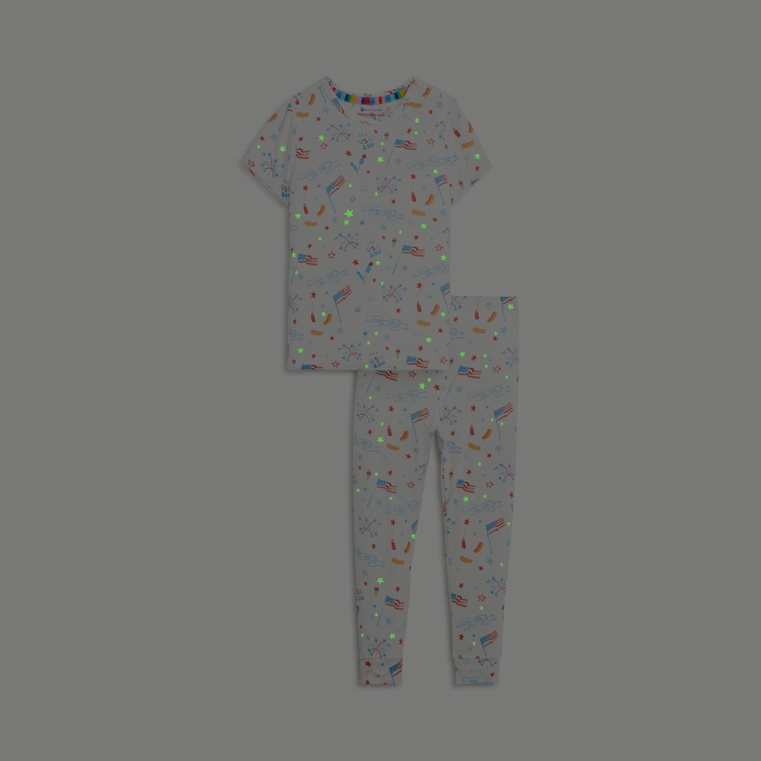Glow in the dark effect in A two-piece white pajama set with a short-sleeve top and shorts featuring a colorful celebratory print of rockets and "1776-2026" banners, by Magnetic Me.