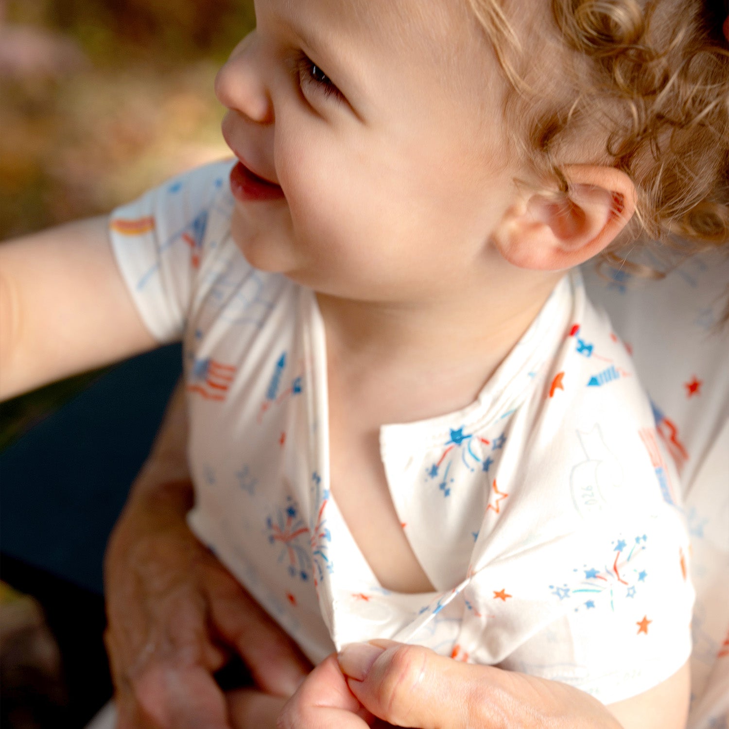 Child sitting outdoors and adult putting the magnet together of A two-piece white pajama set with a short-sleeve top and shorts featuring a colorful celebratory print of rockets and "1776-2026" banners, by Magnetic Me.