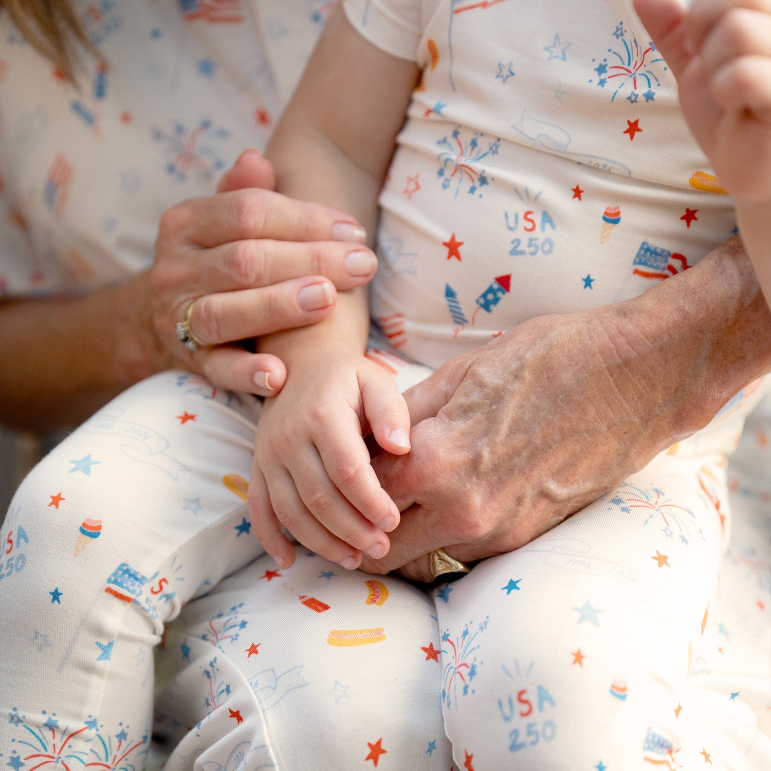 Close-up of a child's hand held by an adult and another person, all wearing A two-piece white pajama sets with a short-sleeve top and shorts featuring a colorful celebratory print of rockets and "1776-2026" banners, by Magnetic Me.