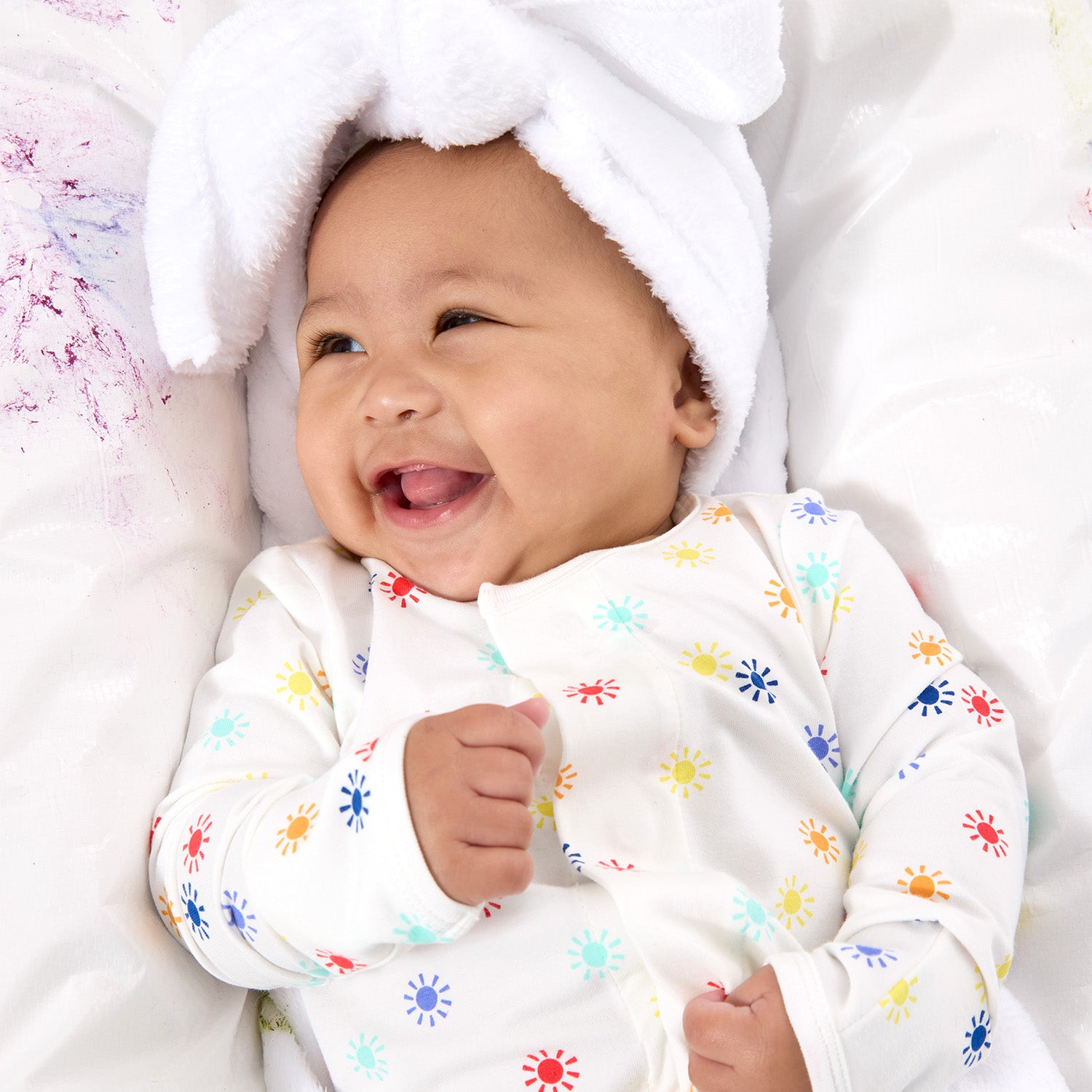 smiling baby on a white surface with abstract paint splashes wearing a multi colored sun print footie by Magnetic Me.