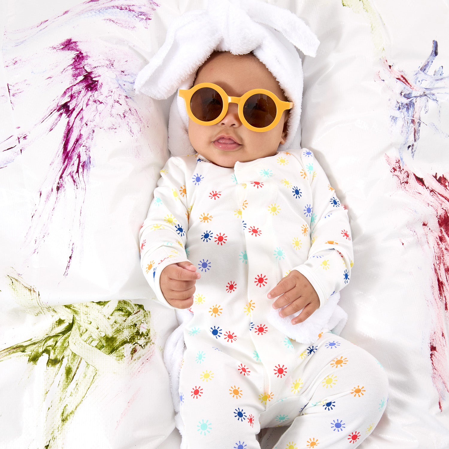 Baby on white surface wearing yellow glasses and  with abstract paint splashes wearing a multi colored sun print footie by Magnetic Me.