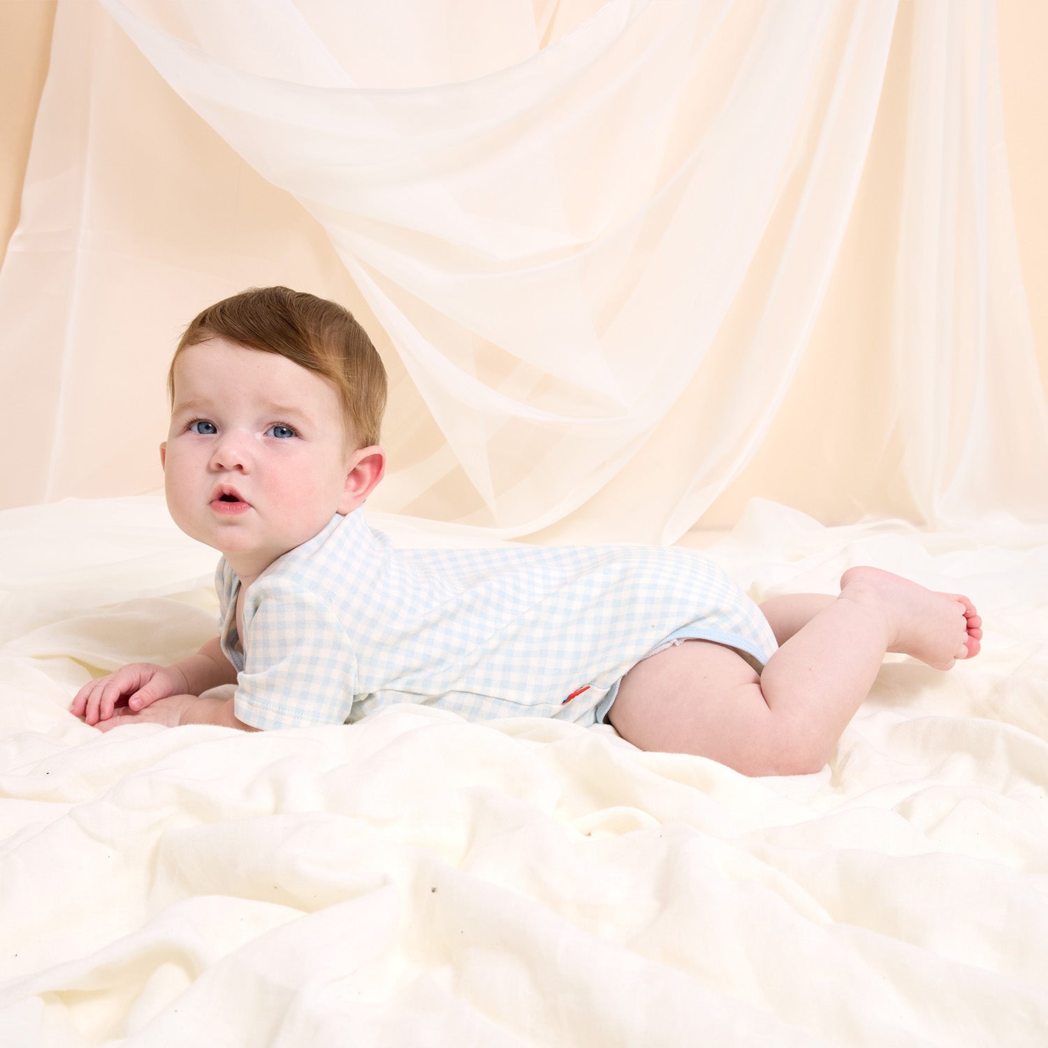 Baby lying on a white fluffy surface wearing a light blue gingham bodysuit, by Magnetic Me.