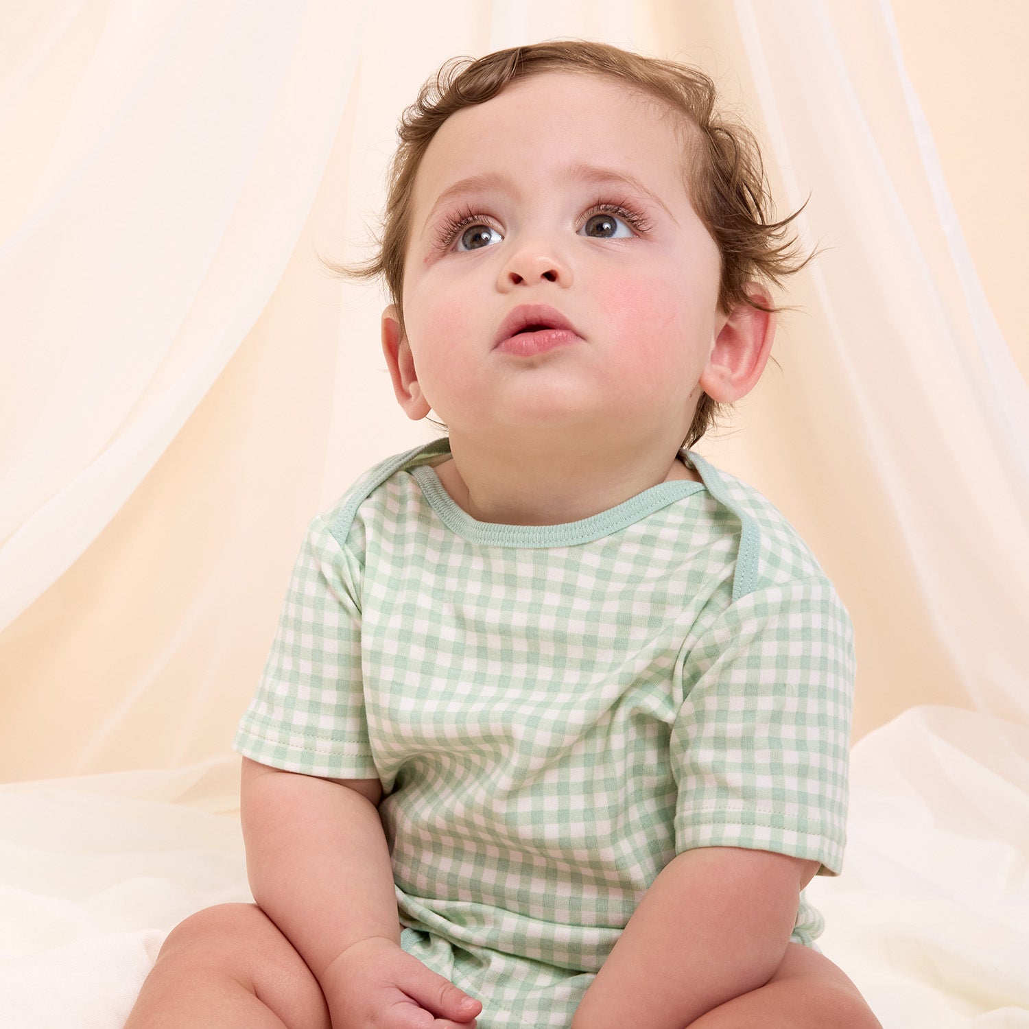 Baby wearing a green checkered bodysuit sitting in front of cream sheets.