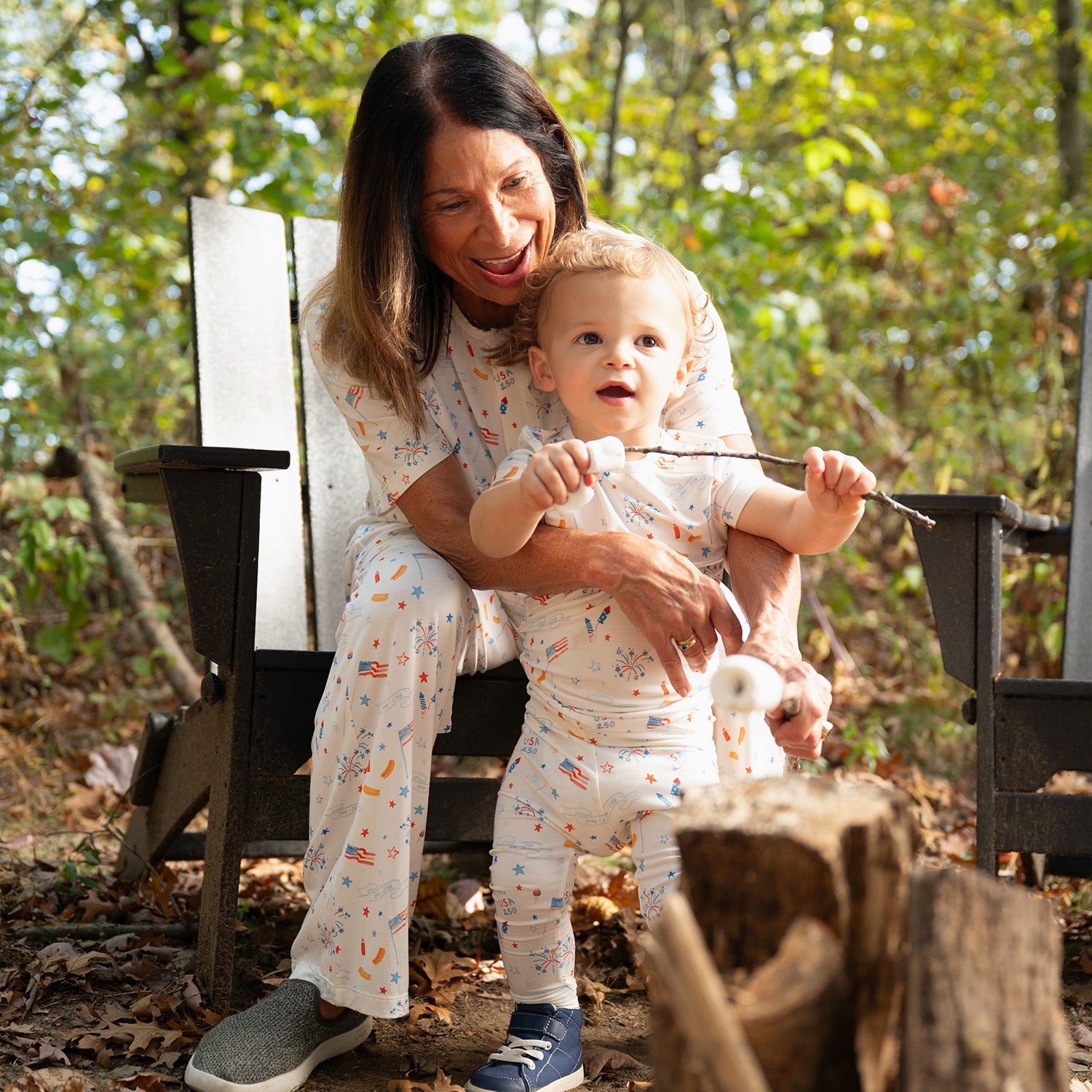Woman and child sitting on a wooden bench outdoors with sticks wearing A two-piece white pajama sets with a short-sleeve top and shorts featuring a colorful celebratory print of rockets and "1776-2026" banners, by Magnetic Me.