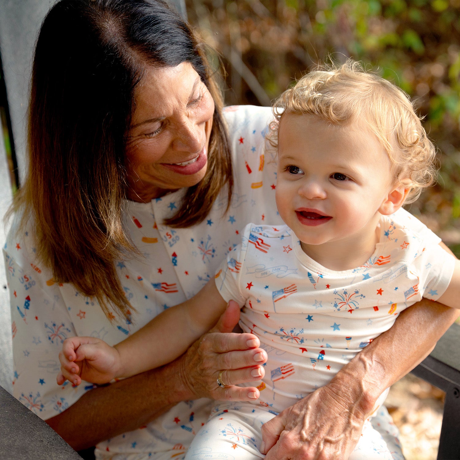 Woman holding a baby both wearing matching  PJ two-piece set featuring a  top and long pants in white fabric with an all-over patriotic holiday print, by Magnetic Me.