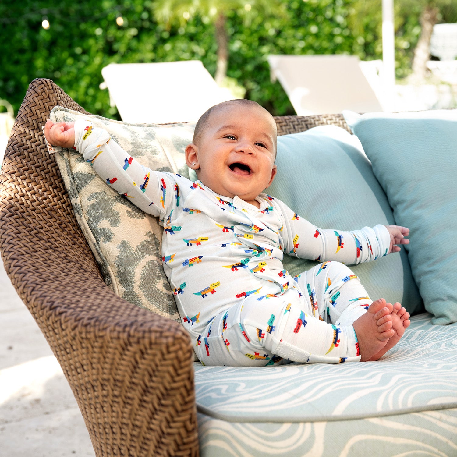 Baby on a cushioned chair outdoors wearing A long-sleeve convertible coverall in light blue featuring an all-over print of colorful, stylized crocodiles and ribbed cuffs, by Magnetic Me.