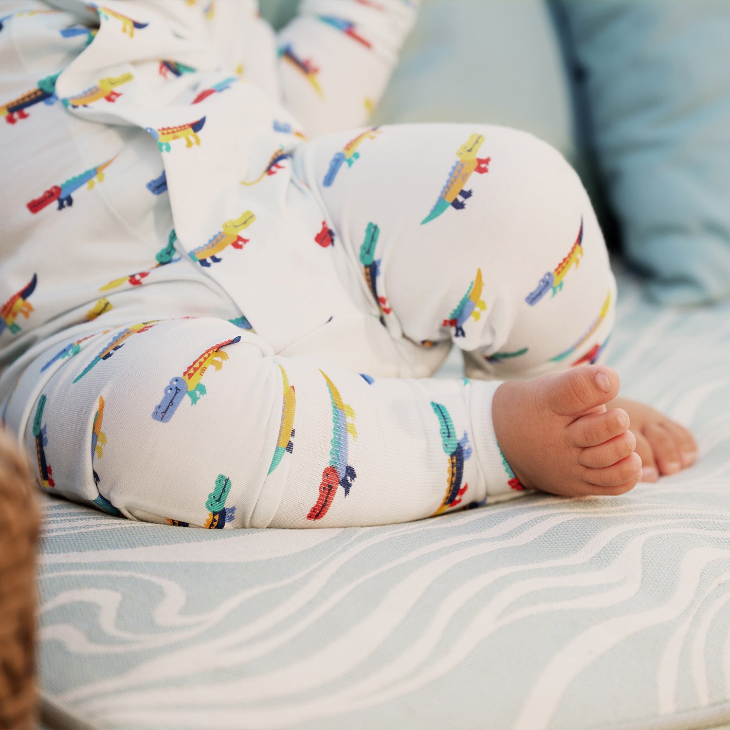 Close up of the babies feet and baby is wearing A long-sleeve convertible coverall in light blue featuring an all-over print of colorful, stylized crocodiles and ribbed cuffs, by Magnetic Me.