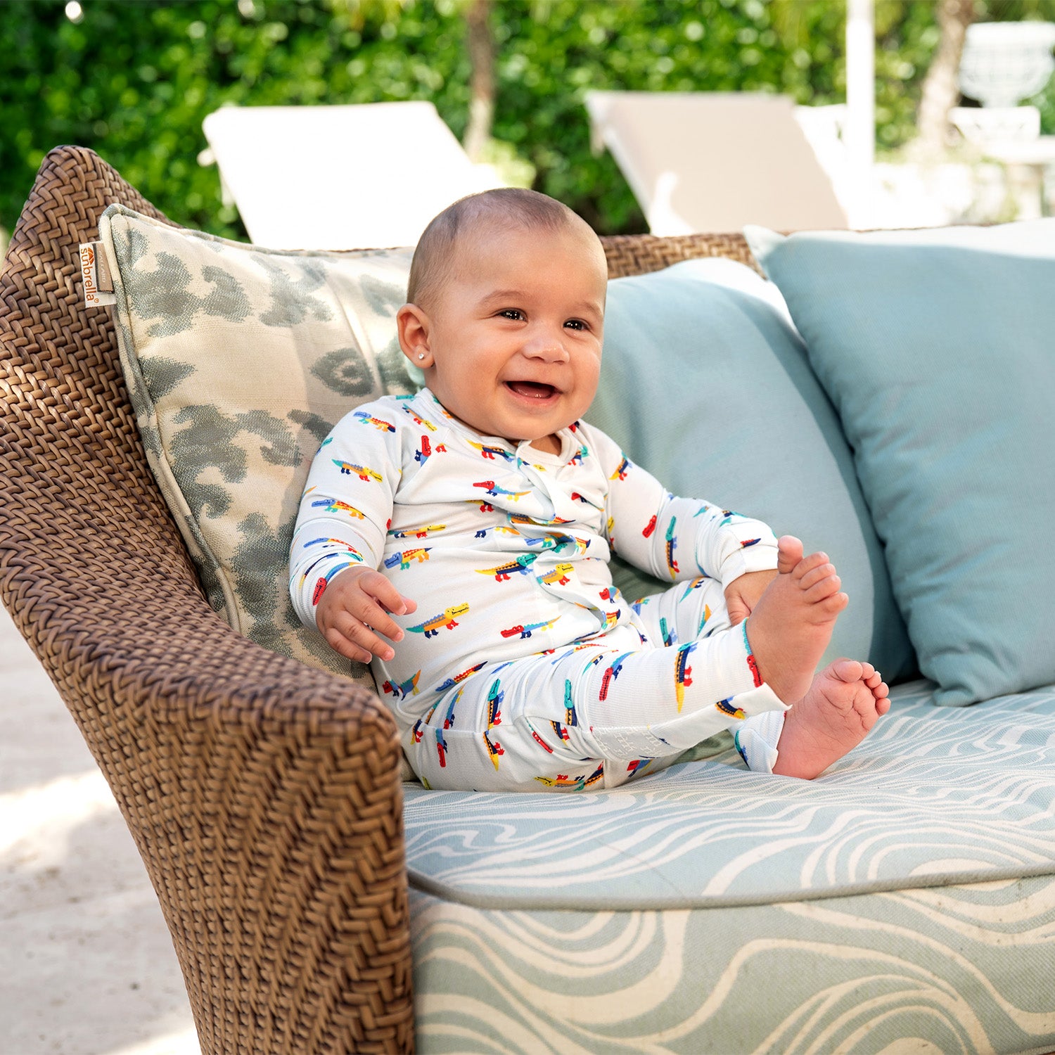 Baby sitting on a cushioned chair outdoors with greenery in the background wearing A long-sleeve convertible coverall in light blue featuring an all-over print of colorful, stylized crocodiles and ribbed cuffs, by Magnetic Me.