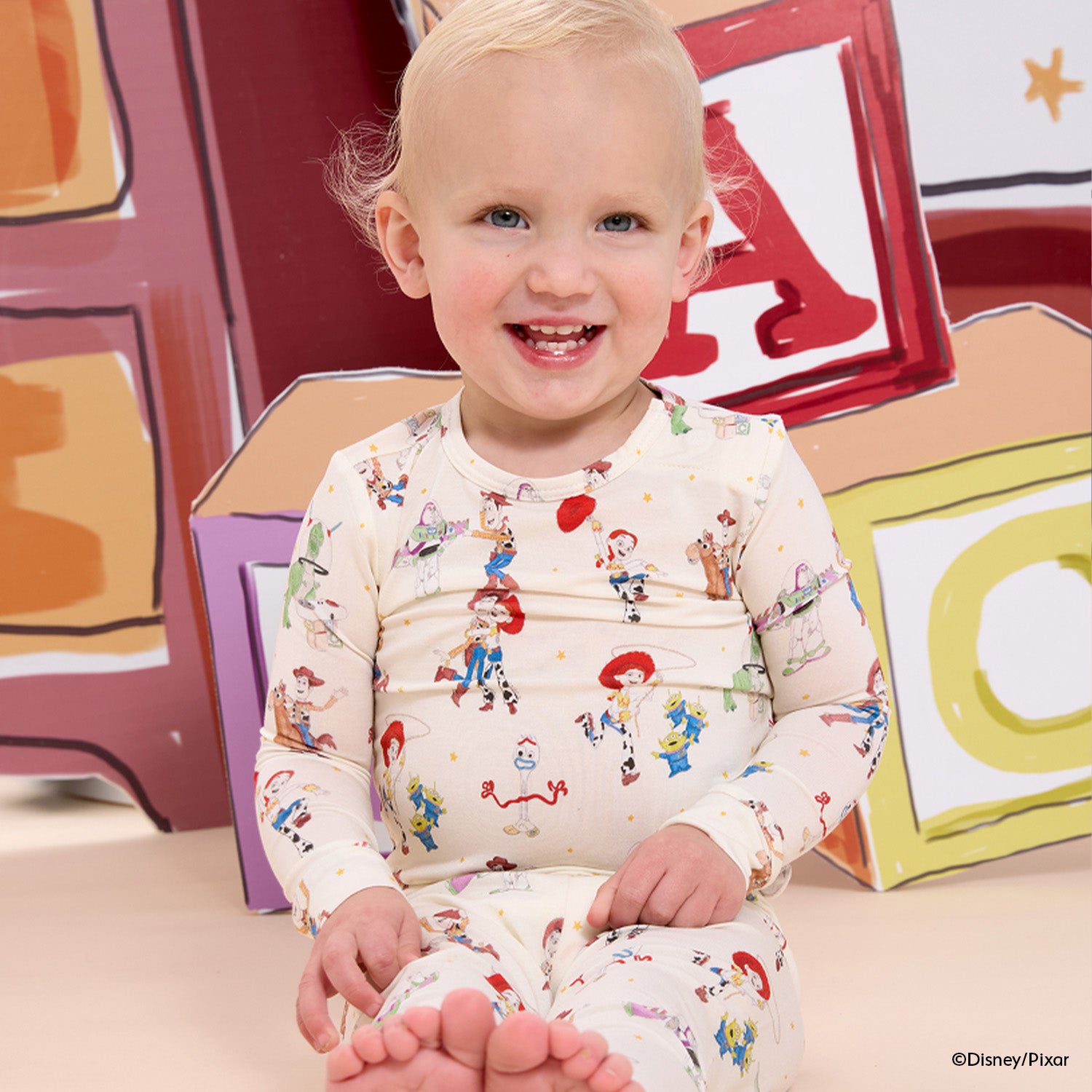 Child sitting in front of colorful letter blocks wearing A two-piece pajama set with a long-sleeve top and matching pants in a cream fabric featuring an all-over Disney Pixar Toy Story print of Woody, Buzz Lightyear, Jessie, Bullseye, Rex, Hamm, Slinky Dog, and the Aliens, by Magnetic Me.