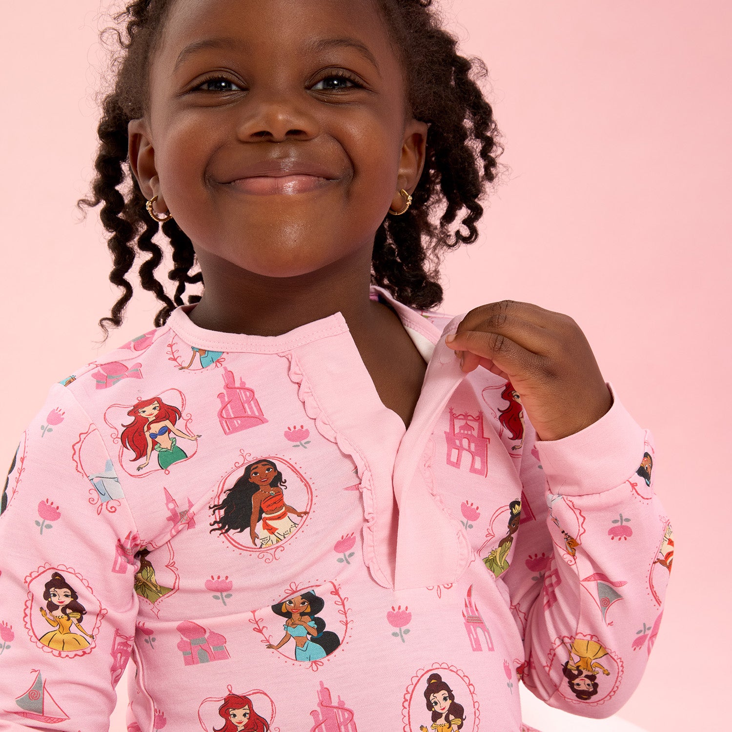 Girl opening the magnet of A two-piece pink pajama set with a ruffled collar top and matching pants featuring an all-over print of Disney Princesses Ariel, Belle, Moana, Tiana, Jasmine, and Cinderella, by Magnetic Me.