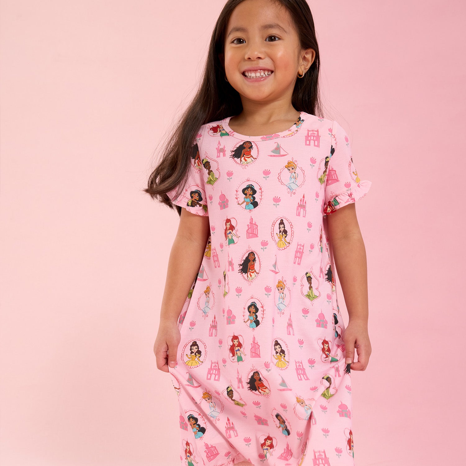 Young girl wearing A pink short-sleeve nightgown with flutter sleeves and a ruffled hem featuring an all-over print of Disney Princesses Ariel, Belle, Moana, Tiana, Jasmine, and Cinderella, by Magnetic Me.