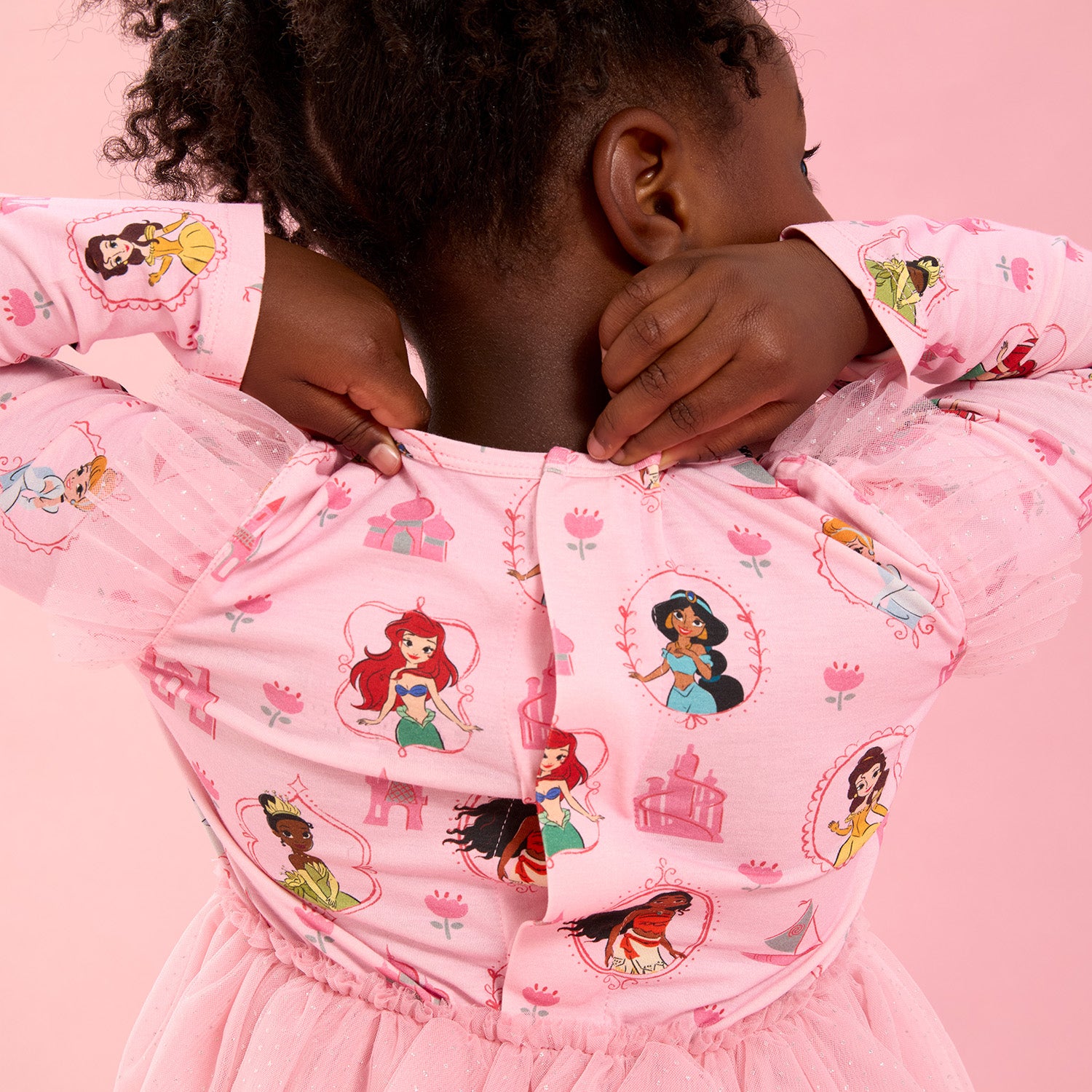 Young girl closing the magnet of A pink short-sleeve nightgown with flutter sleeves and a ruffled hem featuring an all-over print of Disney Princesses Ariel, Belle, Moana, Tiana, Jasmine, and Cinderella, by Magnetic Me.