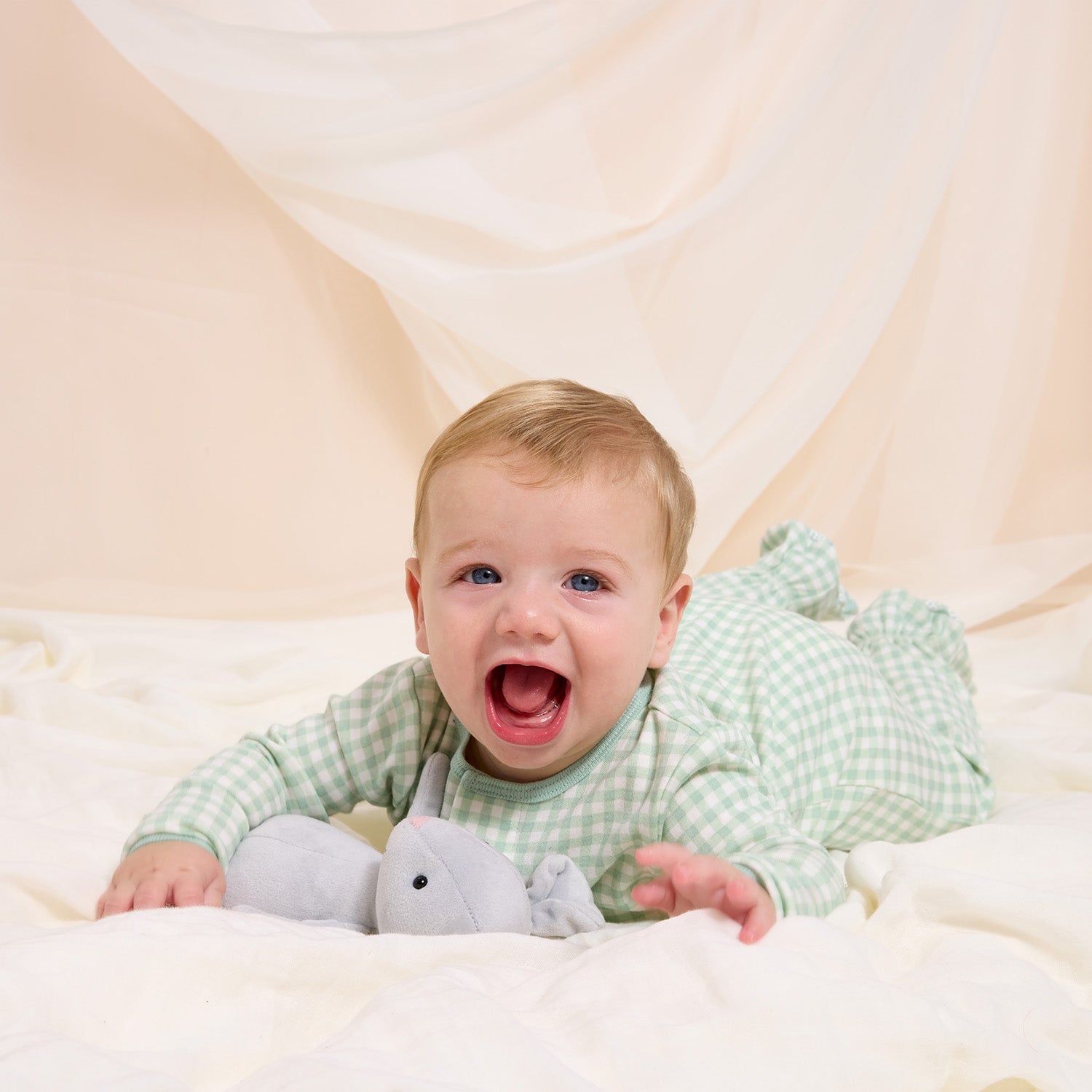 Baby in a light green gingham footie lying on a white blanket with a plush toy, against a beige background by Magnetic Me.