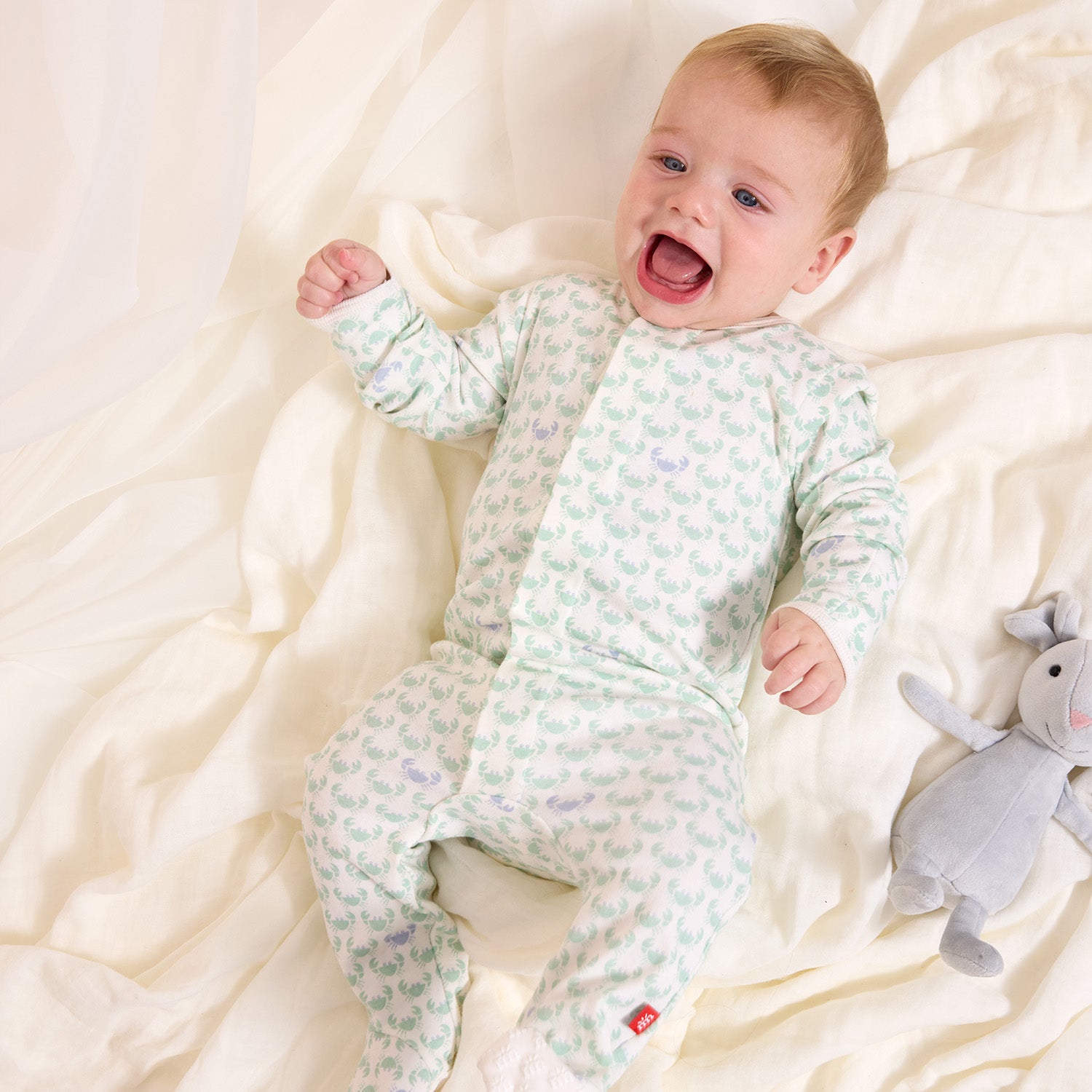 Baby in a light green crap patterned footie lying on a white blanket with a gray toy elephant by Magnetic Me