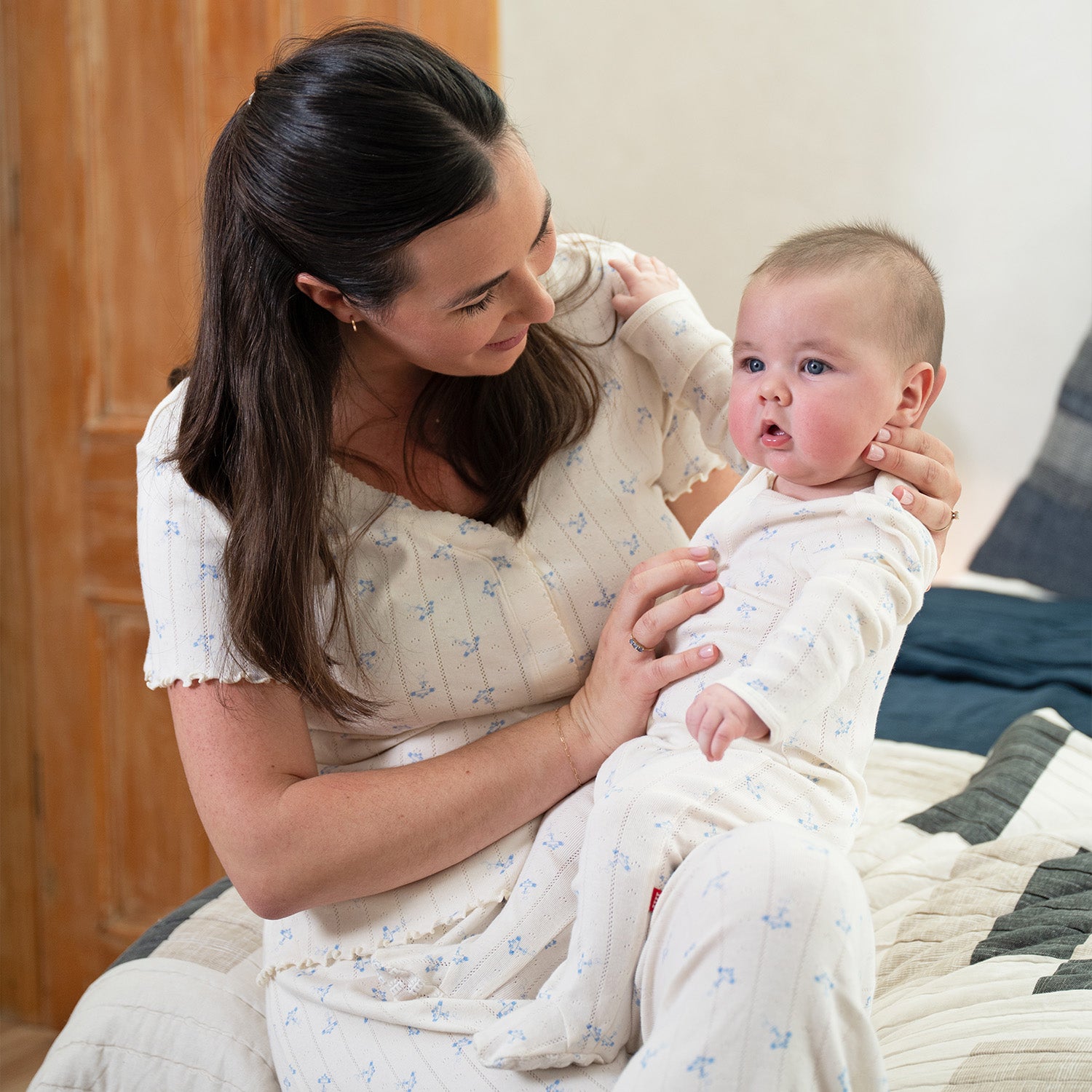 Woman holding a baby wearing A cream-colored pointelle long-sleeve footie with a subtle blue airplane pattern and magnetic closures, by Magnetic Me.