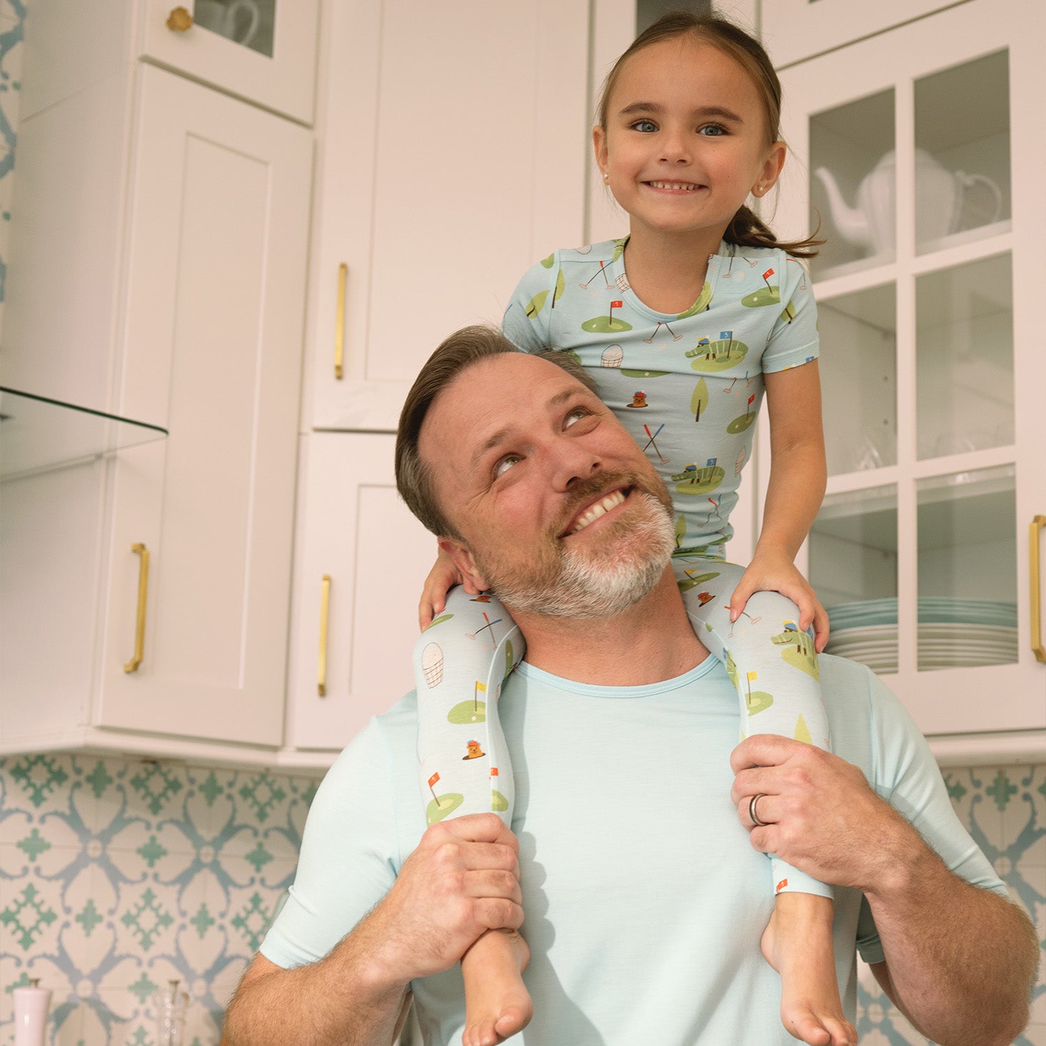 Man and child on his shoulder in matching light blue pajama set with green golf patterned print in a kitchen