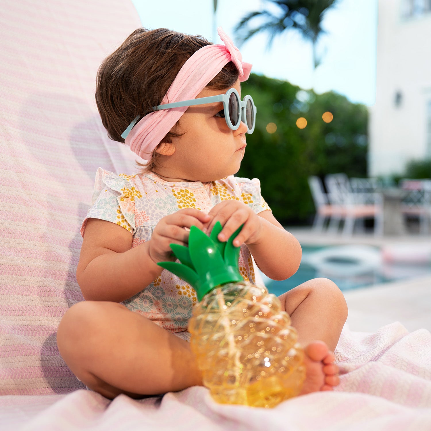 Baby holding a pineapple-shaped cup outdoors while wearing A short-sleeve baby romper with ruffled shoulders and elastic leg openings featuring a dense pastel floral print of pink, yellow, and light blue wildflowers on a white background, by Magnetic Me.