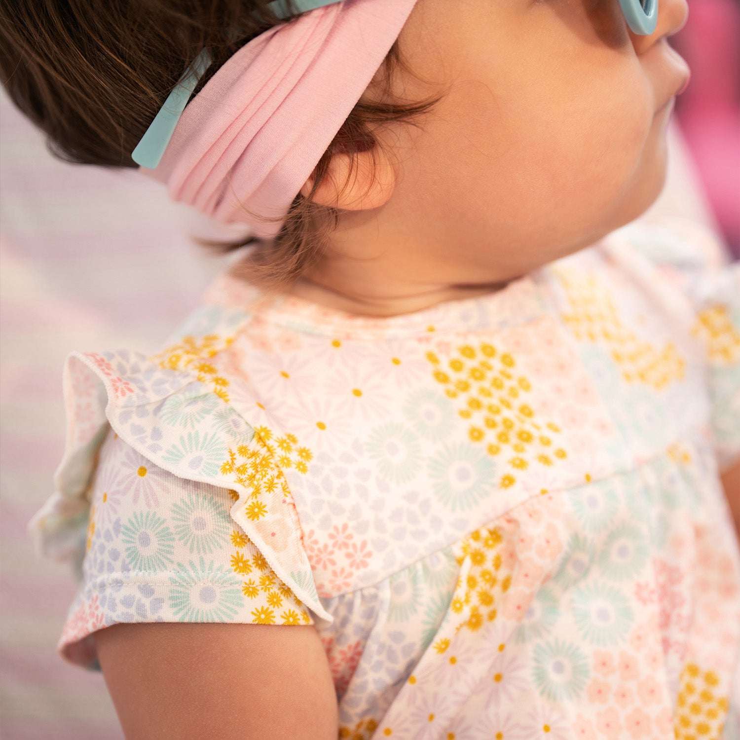 Close-up of a child wearing a pink headband and A short-sleeve baby romper with ruffled shoulders and elastic leg openings featuring a dense pastel floral print of pink, yellow, and light blue wildflowers on a white background, by Magnetic Me.