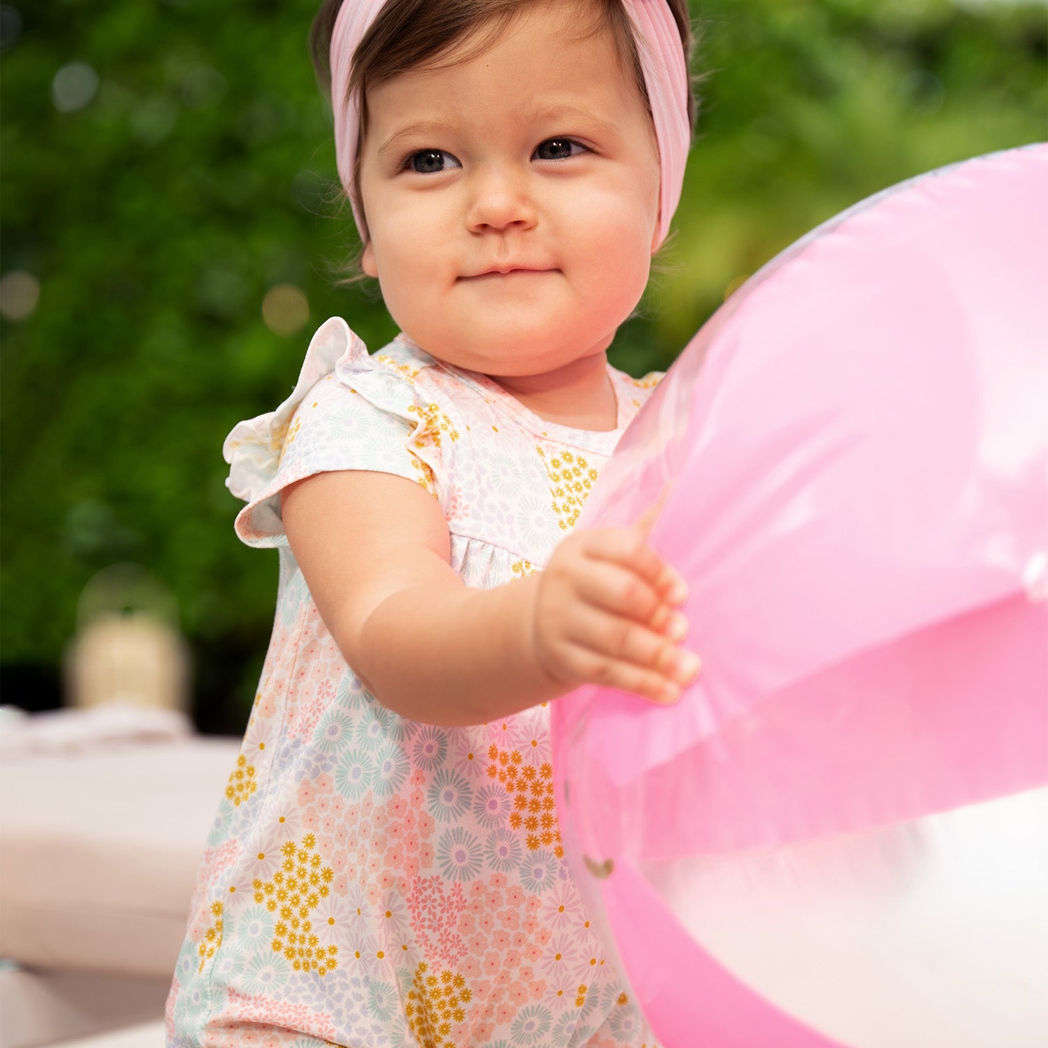 Baby holding a pink balloon outdoors wearing  A short-sleeve baby romper with ruffled shoulders and elastic leg openings featuring a dense pastel floral print of pink, yellow, and light blue wildflowers on a white background, by Magnetic Me.